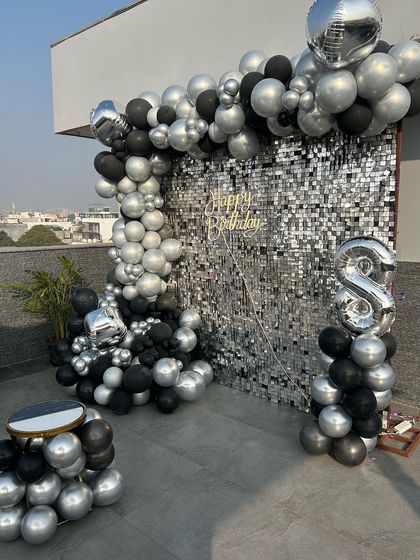 A complete birthday photo corner on a terrace, featuring a silver sequin wall, a black and silver balloon arrangement, and a small table for the cake.