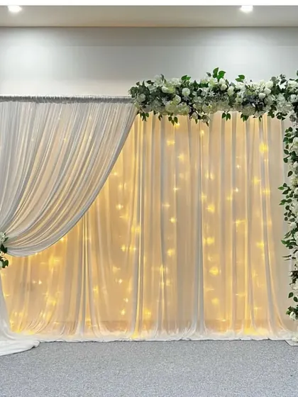 A classic and sophisticated backdrop made of white drapery and fairy lights. The look is completed with a beautiful white and green floral garland framing the top and side, perfect for an elegant celebration.