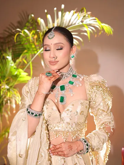 A full view of the soft glam morning bridal look. The ivory and gold lehenga paired with emerald jewelry is complemented by makeup that is both fresh and regal.