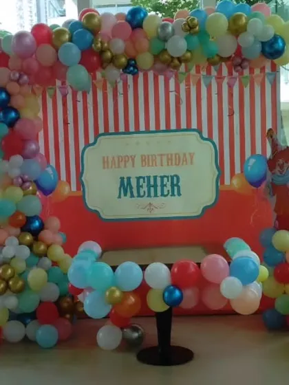 A cheerful carnival-themed birthday setup for Meher. This backdrop features classic red and white stripes, a personalized name sign, and a vibrant, multi-colored organic balloon garland around the cake table.