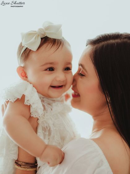 A beautiful close-up of a mother and her smiling baby girl. The baby's lacy white outfit and bow add to the sweetness of this portrait.