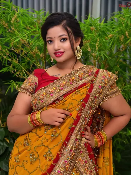 A three-quarter length portrait against a simple background with green foliage. This shot highlights the intricate embroidery of the yellow and red saree.