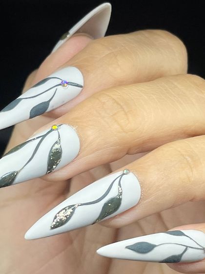 A sophisticated design on long stiletto nails, featuring hand-painted leaves on a matte white base with small, sparkling gems.