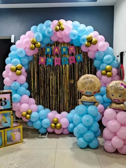 A classic baby shower decoration with a circular balloon arch in pink and blue, a gold shimmer curtain, and foil baby balloons. The transparent 'BABY' blocks are filled with matching balloons for a cohesive look.