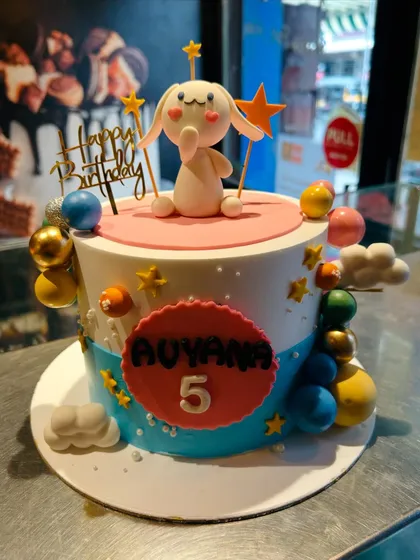 A whimsical birthday cake featuring a cute white bunny character amidst colorful planets and stars.