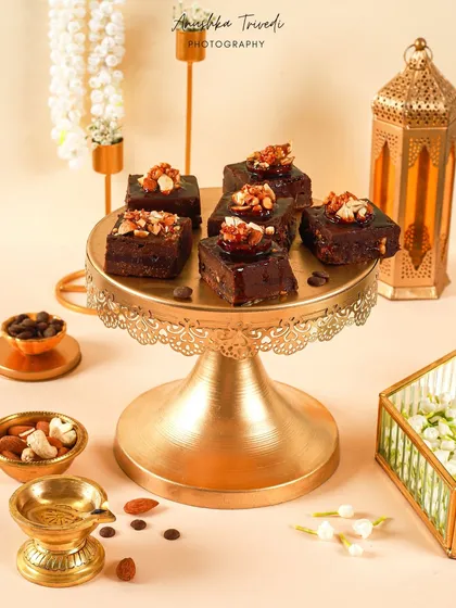 A platter of decadent, nut-topped brownies on a golden stand. The styling with a diya and lantern gives it a warm, festive glow perfect for Diwali.