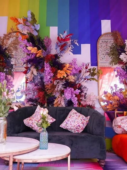 A bold and artistic seating area from a colorful Haldi event. The rainbow-hued backdrop is complemented by lush, wild floral arrangements and a mix of modern and traditional furniture, creating a unique and vibrant photo corner.