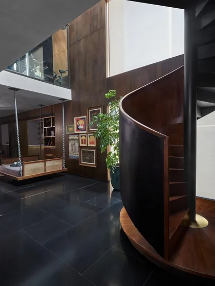 The spiral staircase at the Baid Residence, viewed from the lower level. The space combines modern architectural elements with traditional touches like a custom wooden swing and a gallery wall of Indian art.