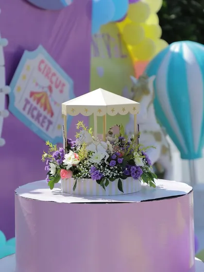 A detailed shot of a miniature carousel centerpiece on a tiered stand. The fresh flowers and hot air balloon prop in the background show the cohesive event design.