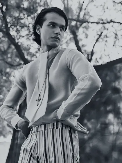 A black and white editorial shot for Kaltblut Magazine. The androgynous styling, featuring a sheer shirt with a built-in scarf, striped trousers, and a cross pendant, explores themes of gender fluidity in high fashion.
