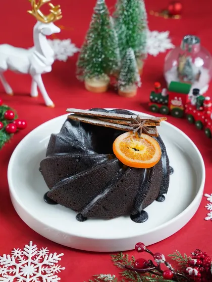 A close-up of a dark chocolate bundt cake, garnished with a dried orange slice and cinnamon, on a festive red background. The simple composition highlights the dessert's elegant details.
