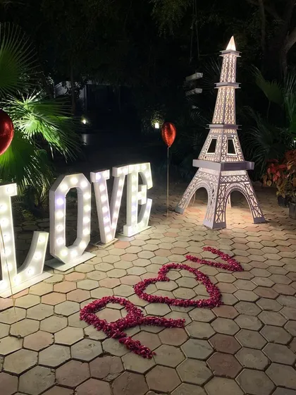 A detailed look at our unique prop combination for a romantic declaration. The setup includes large, bright "LOVE" marquee letters, an illuminated Eiffel Tower model, and the words "I Love You" spelled out with rose petals on the ground.