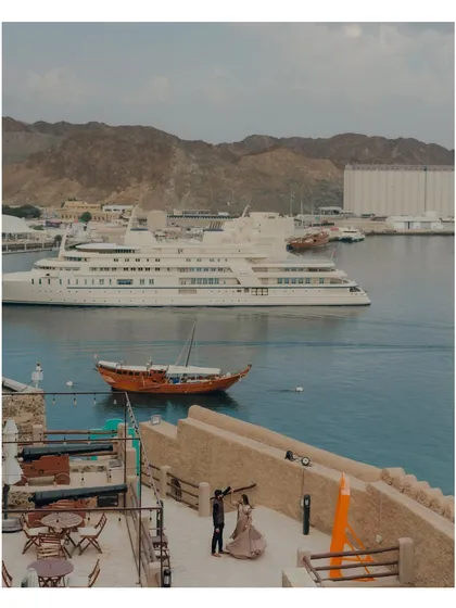 A wide shot capturing the scale of the Muscat harbour. The couple provides a focal point on a terrace overlooking the water, with luxury yachts and traditional dhows in the background.