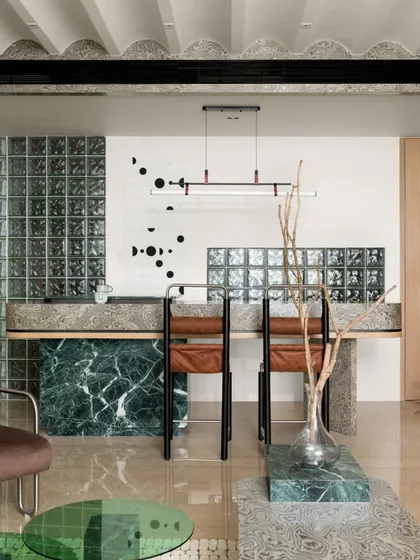 A view of the open-plan living and dining area, showcasing the interplay of materials. The green marble breakfast bar, glass block partitions, and warm wooden cabinetry create a harmonious flow between the different zones of the home.