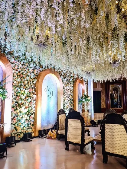 A close-up of the engagement ceremony stage, showing the wall of flowers, the elegant monogram, and the ceiling of cascading white wisteria, creating a lush and romantic atmosphere.