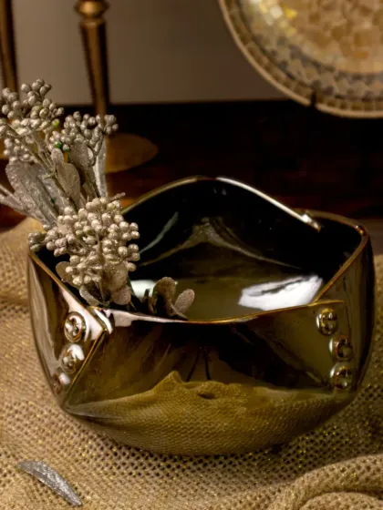 The unique, folded design of this glossy metallic bowl is highlighted with shimmering silver foliage. The rustic jute background provides a textural contrast to the bowl's smooth, elegant finish.