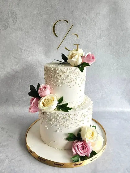 Simple, elegant, and full of grace. This two-tiered wedding cake is decorated with delicate pearl-like sprinkles and adorned with fresh pink and white roses. The custom initial topper adds a perfectly personal touch.