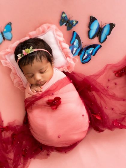 A sweet newborn girl swaddled in pink, surrounded by delicate blue butterflies against a soft pink background. This is a popular fantasy theme for baby girls.