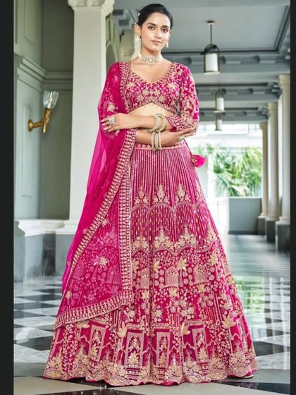 A full-length view of a stunning fuchsia pink bridal lehenga. The entire outfit is covered in intricate gold embroidery, creating a luxurious and celebratory look perfect for a wedding.