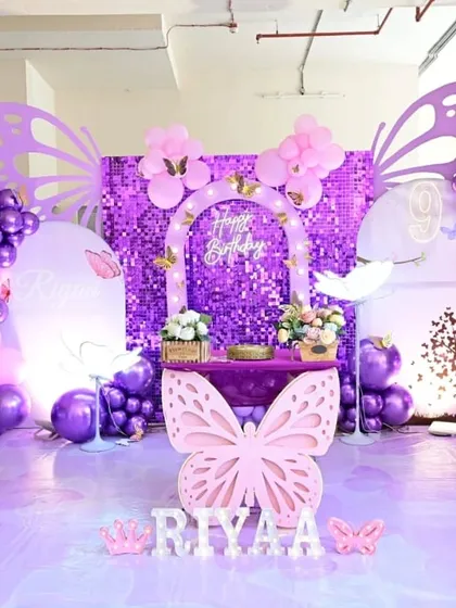 A butterfly theme party featuring a stunning purple sequin wall for a shimmering effect. The setup includes custom butterfly wing cutouts, a themed dessert table, and personalized marquee letters.