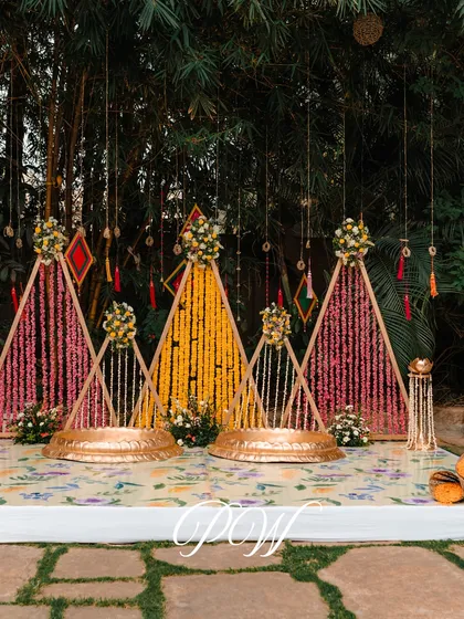 A unique Haldi backdrop made of triangular frames woven with pink and yellow flowers, creating a modern and artistic look.