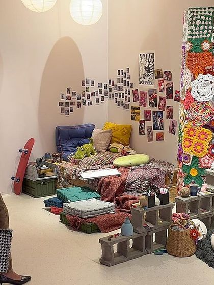 A visitor explores the 'Goblin Mode' installation, a space designed for comfort and self-indulgence. The environment is a tactile and sensorial landscape, filled with hand-tufted textures, crochet forms, and personal objects.