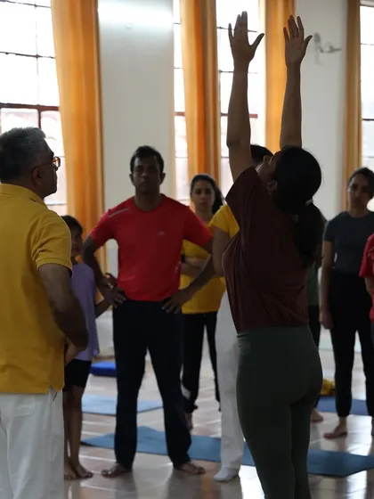 A teacher guides a student in the raised arms pose, the second step of Surya Namaskar. Our classes focus on step by step learning for a strong foundation.