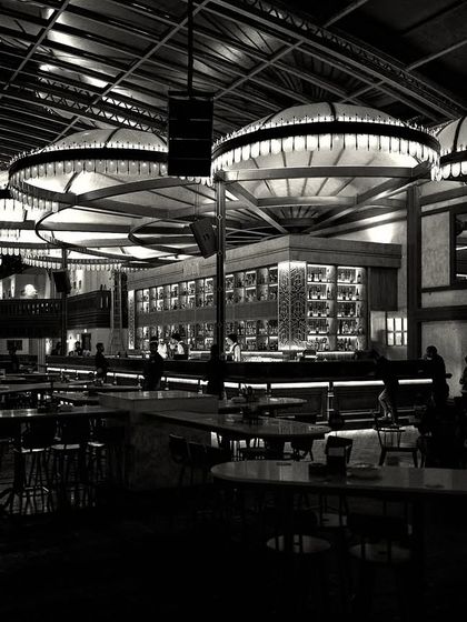 A black and white photograph capturing the dramatic lighting of the main bar at night. The monochrome treatment emphasizes the form and luminosity of the custom fixtures.