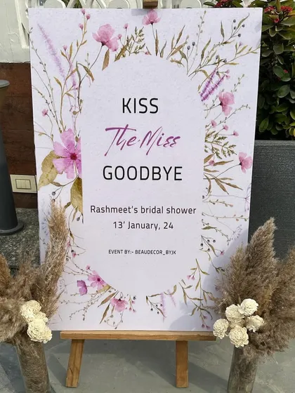 A charming welcome sign for a bridal shower that reads "Kiss The Miss Goodbye," framed by rustic pampas grass arrangements.