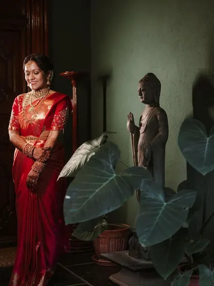 A bride in a red saree, standing by a Buddha statue, a portrait that feels both serene and vibrant.