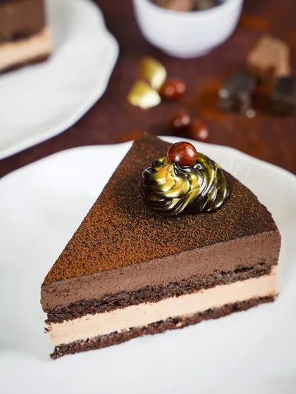 A slice of rich, multi-layered chocolate and hazelnut mousse cake. The clean, bright photography style focuses on the different textures and layers of the decadent pastry.