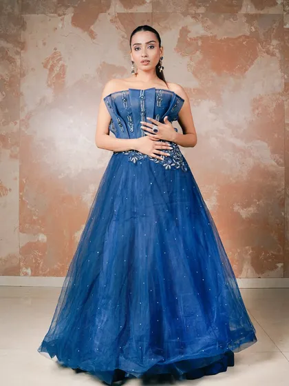 A full-length portrait of a model in a royal blue off-shoulder ball gown. The simple, textured background ensures that the focus remains entirely on the dress's stunning color, structured corset, and voluminous tulle skirt.