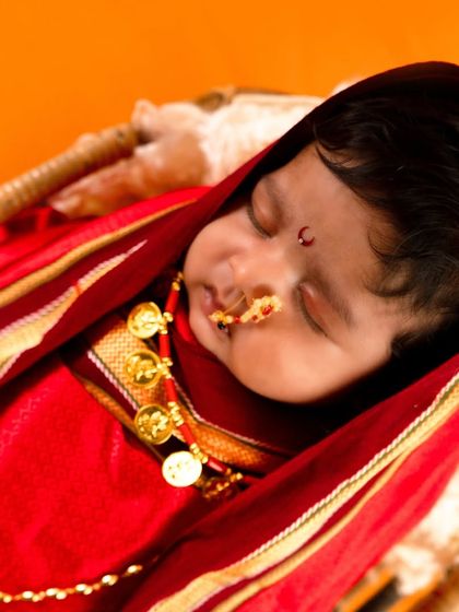 A close-up of a newborn in a traditional Maharashtrian look, dressed in a red nauvari-style wrap with a classic nose ring and bindi.