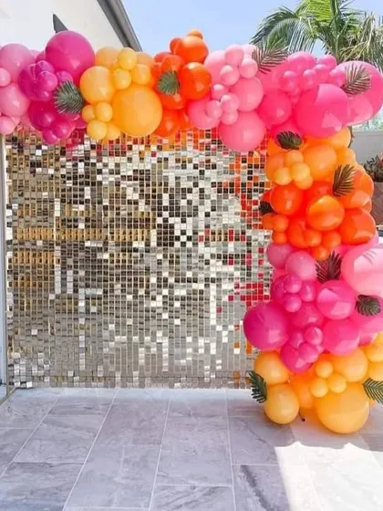 A vibrant balloon arch against a gold sequin wall, perfect for an outdoor poolside or patio party. The tropical colors of pink, orange, and yellow are accented with palm leaves.