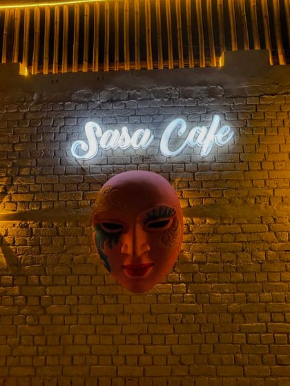 Our iconic "Sasa Cafe" neon sign against a rustic brick wall, with a decorative mask adding a touch of mystery. This is one of our most popular photo spots.