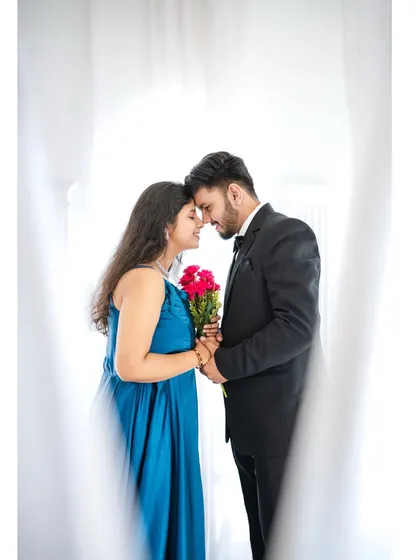 A soft, romantic close-up of a couple framed by white curtains, with the bride holding a bouquet of red roses. This intimate portrait highlights the couple's connection amidst a dreamy studio setting.
