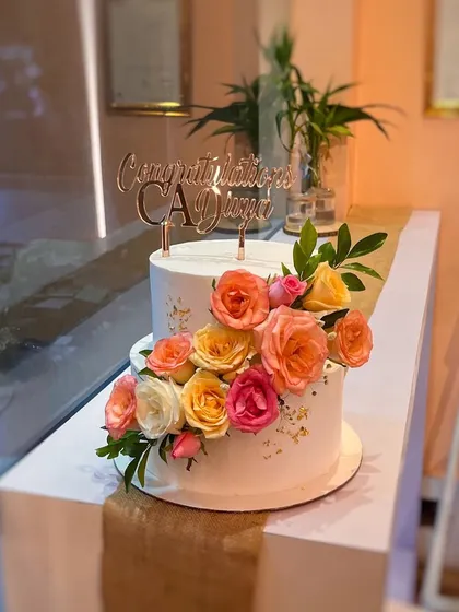 We make cakes for every celebration, including this one for a new Chartered Accountant. This two-tier white cake is decorated with a vibrant cascade of fresh, colorful roses and a custom "Congratulations CA" topper.