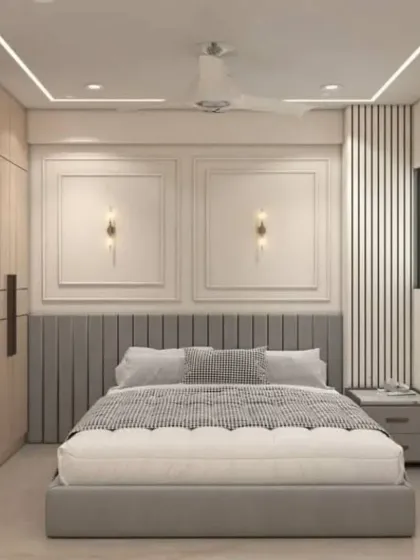 This bedroom showcases classic elegance with modern touches. The grey upholstered bed is set against a wall with decorative molding and stylish sconces, while the cove lighting adds a soft glow.