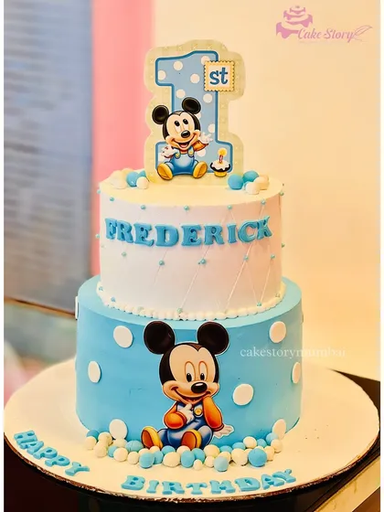 A classic Mickey Mouse first birthday cake. This two-tier design in blue and white is decorated with polka dots and adorable baby Mickey figures.