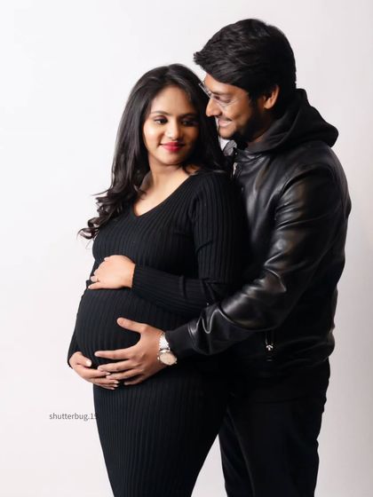 A sweet and simple couple's portrait against a clean white background. Their casual black outfits and the gentle way they hold the baby bump make for a warm and modern maternity photo.