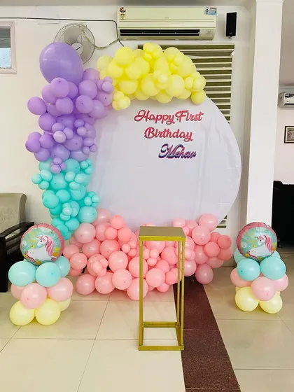 A simple and colorful pastel decoration for a first birthday party. The circular backdrop can be customized with the baby's name, and the organic balloon garland uses a mix of soft, cheerful colors.