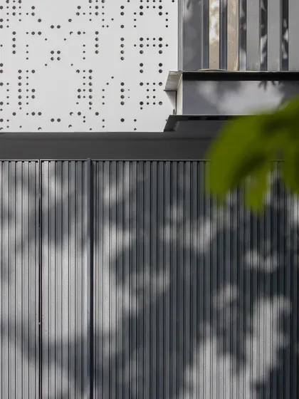 A detail shot focusing on the texture of the fluted gate panels against the perforated jali. The shadows from the leaves add a soft, organic element to the sharp, geometric design.