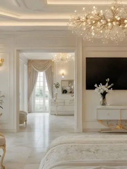 An all-white luxury bedroom that exudes Parisian chic. The room features ornate wall molding, a stunning crystal chandelier, and a view into an equally elegant living space.