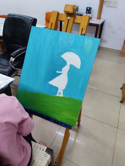 A student works on an imaginative painting of a girl with an umbrella in a field, using a palette knife and brush to create texture.