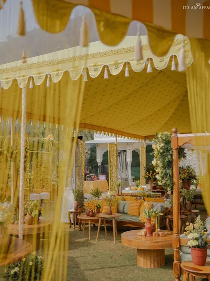A view through the sheer yellow curtains of a tent, revealing the lush and festive Haldi setup inside.