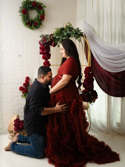A classic and romantic maternity pose, with the husband-to-be kneeling to kiss the baby bump. The flowing red gown and floral setting add to the fairy-tale feel of the portrait.