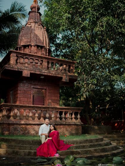 A romantic portrait of the couple seated at the base of a temple, with the bride's red saree spread out. The scene is peaceful, traditional, and full of grace.