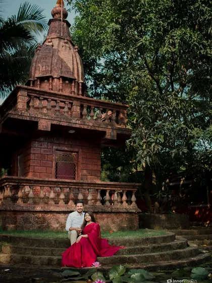 A romantic portrait of the couple seated at the base of a temple, with the bride's red saree spread out. The scene is peaceful, traditional, and full of grace.