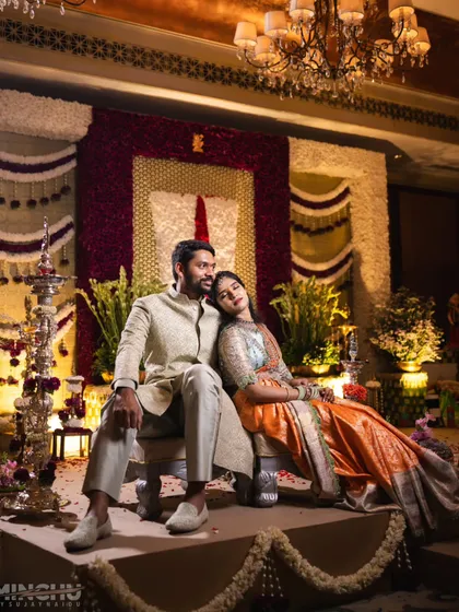 A quiet moment during a traditional Muhurtham ceremony. The backdrop is intricately designed with floral panels, including a prominent Tirunamam symbol made of red and white flowers, blending sacred tradition with beautiful aesthetics.
