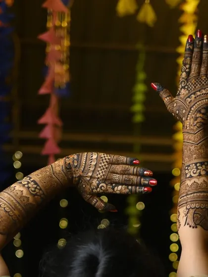 A beautiful shot of a bride with her hands raised, showing the back of her arm mehendi against a festive background. The design is both bold and graceful.